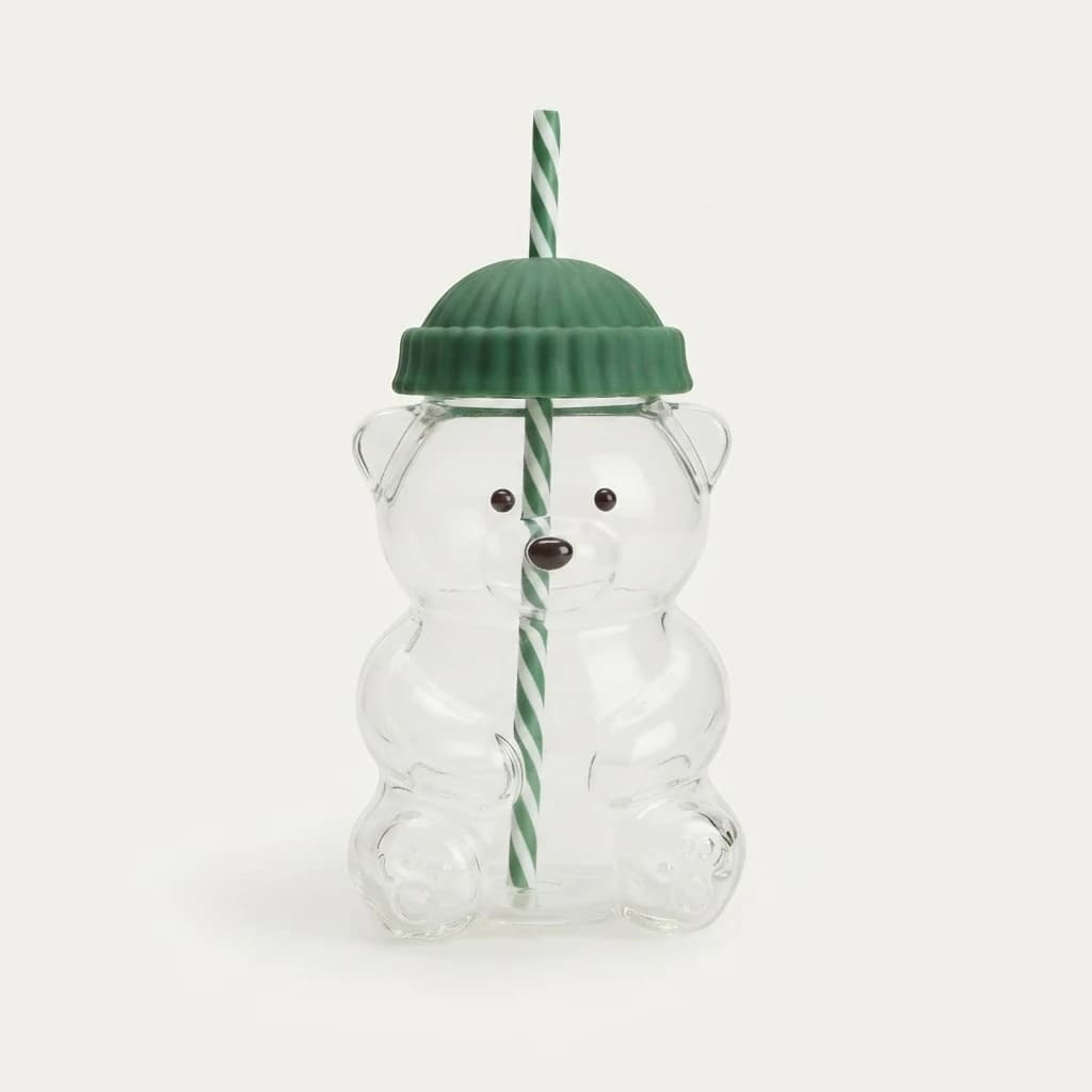 Clear glass bear tumbler standing upright with green beanie lid and striped straw on plain ground.