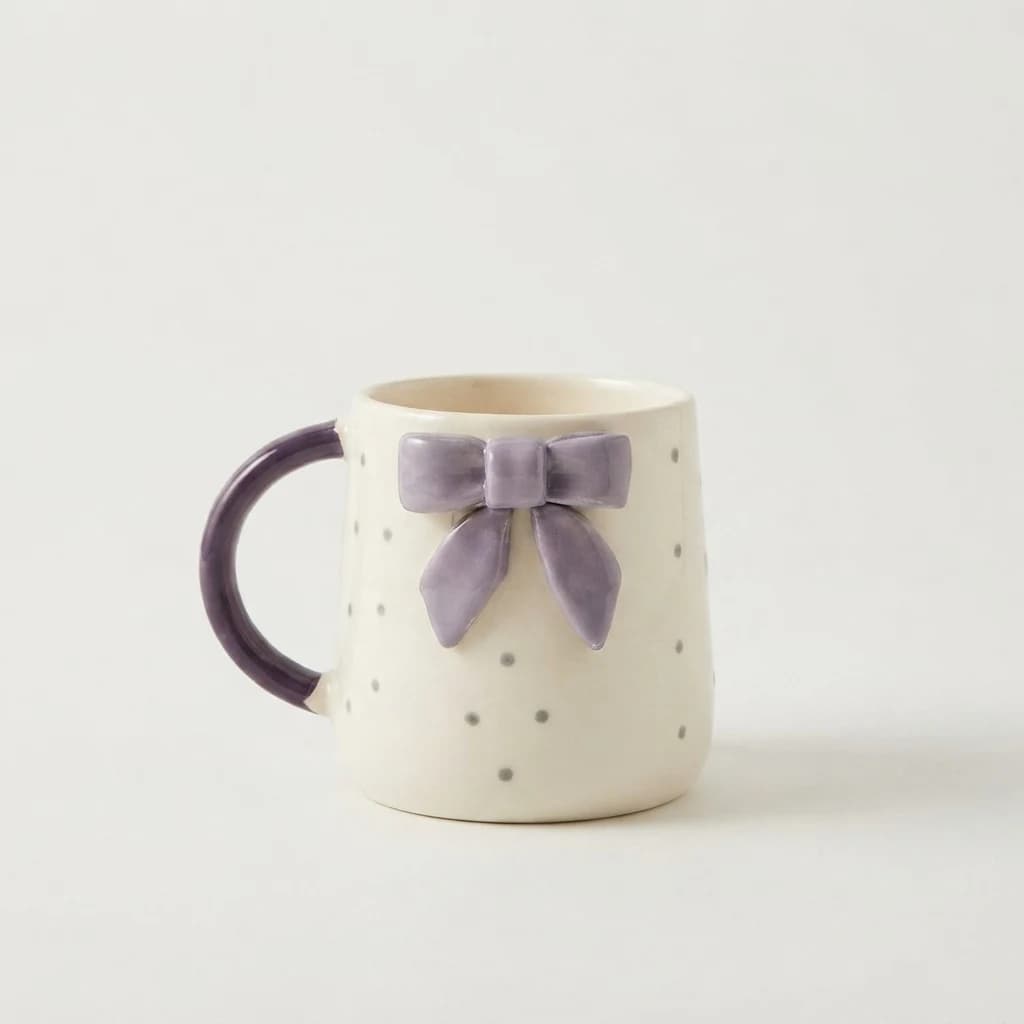 Off-white ceramic mug featuring a raised purple bow and tiny grey polka dots.